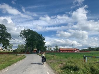 Radfahrer mit Packtaschen auf asphaltierter Landstraße zu roten Bauernhäusern. Grüne Felder und Bäume unter blauem Himmel mit weißen Wolken.