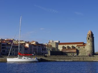 Weißes Segelboot im Hafen von Collioure mit historischem Kirchturm und Ufergebäuden unter blauem Himmel in den Pyrenäen.