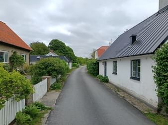 Ruhige Asphaltstraße durch ein schwedisches Dorf mit traditionellen Häusern auf beiden Seiten, grünen Gärten und bewölktem Himmel am Österlenleden.