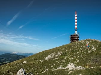 Auf einer Bergspitze steht ein Rot-Weiss gestreifter Funkmast vor einem strahlend blauen Himmel. Ganz klein versteckt sieht man 2 Wandere die den Berg hinunter laufen.