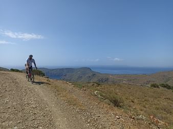 Radfahrer auf einem Schotterweg in Nordkatalonien mit Mittelmeer und Hügellandschaft im Hintergrund unter klarem blauen Himmel.