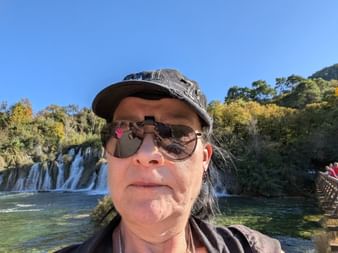 Frau mit Sonnenbrille und Kappe macht Selfie bei den Krka-Wasserfällen in Norddalmatien. Kaskadierende Wasserfälle und grüne Vegetation im Hintergrund.