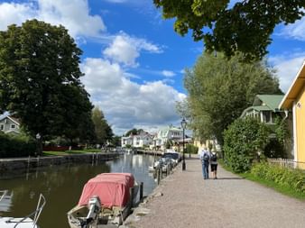 Zwei Personen spazieren entlang eines Kanalwegs in Trosa mit bunten Holzhäusern, vertäuten Booten und großen Bäumen unter blauem Himmel.