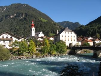 Alpendorf Pfunds in Tirol mit weißem Kirchturm, traditionellen Gebäuden und türkisfarbenem Fluss, umgeben von bewaldeten Bergen.