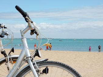 Zwei silberne Fahrräder am Sandstrand mit türkisfarbenem Meer und Strandbesuchern im Hintergrund unter blauem Himmel an der Atlantikküste.