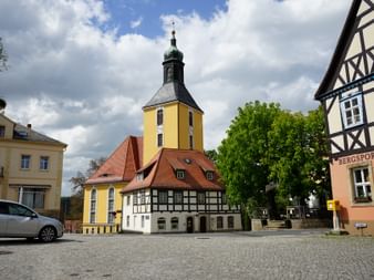 Kopfsteinpflasterplatz in Hohnstein mit gelbem Kirchturm, Fachwerkhäusern und bewölktem Himmel. Ein weißes Auto ist links geparkt.