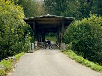 Überdachte Holzbrücke mit Radfahrer neben Velo auf gepflastertem Weg bei Saignelégier, umgeben von grünem Wald und Hügeln.