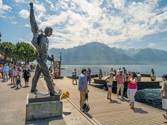 Statue en bronze de Freddie Mercury avec le poing levé sur la promenade de Montreux. Des touristes se rassemblent autour avec le lac Léman et les Alpes.