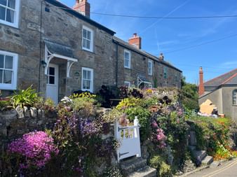 Reihe traditioneller Steinhäuser in Cornwall mit bunten Blumengärten. Weißes Tor führt zu terrassierten Gärten mit rosa, lila Blüten unter blauem Himmel.