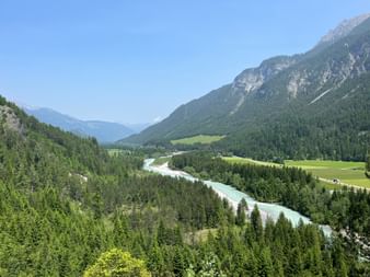 Luftaufnahme des türkisfarbenen Lech, der sich durch ein bewaldetes Alpental mit grünen Wiesen und Bergen unter blauem Himmel schlängelt.