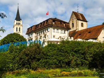 Blick auf Romanshorn mit weißer Kirche mit hohem Turm, historischen Gebäuden mit roten Ziegeldächern und Schweizer Flagge. Grüne Bäume und Gärten im Vordergrund.