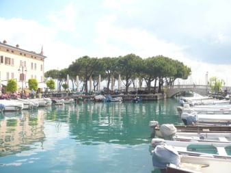 Marina in Desenzano mit weißen Booten im türkisfarbenen Wasser. Historische Gebäude säumen das Ufer mit Fußgängerbrücke und Pinien.