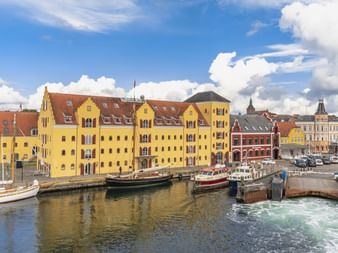 Historischer Hafen von Svendborg mit gelben Ufergebäuden, traditionellen Booten im Hafen und bunten Häusern unter blauem Himmel.