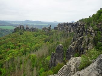 Dramatische Sandsteinfelsen der Schrammsteine erheben sich über dichtem Wald entlang des Malerwegs, mit fernen Bergen unter bewölktem Himmel.