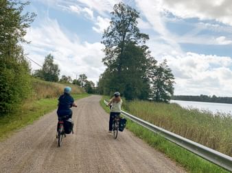 Zwei Radfahrer mit Taschen auf asphaltierter Straße an einem See bei Mariefred, Schweden. Links Bäume, rechts Wasser unter bewölktem Himmel.