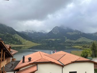 Blick vom Zimmer der Hostellerie Schwarzsee auf den See, umgeben von bewaldeten Bergen unter bewölktem Himmel, mit Ziegeldächern im Vordergrund.