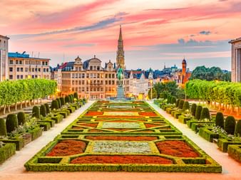 Formale Gärten am Mont des Arts in Brüssel mit bunten Blumenbeeten, geschnittenen Hecken und Stadtsilhouette unter rosa Abendhimmel.