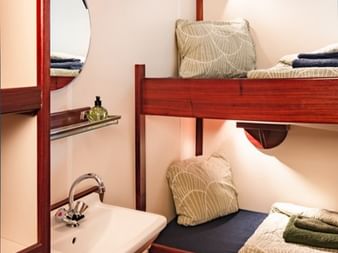Kompakte Schiffskabine auf dem Segelschiff Antigua mit hölzernen Etagenbetten, kleinem Waschbecken, rundem Bullauge und grün gemusterter Bettwäsche.