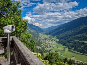 Aussichtsfernrohr auf Holzplattform mit Blick auf das Mölltal mit grünen Wiesen, Dorf und bewaldeten Bergen unter blauem Himmel mit Wolken.
