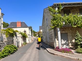 Cycliste en maillot jaune dans une rue étroite de Candes-Saint-Martin avec bâtiments en pierre, végétation verte et fleurs sous ciel bleu.