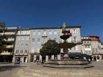 Steinbrunnen mit Statue auf dem Platz in Barcelos, umgeben von traditionellen portugiesischen Gebäuden mit Azulejo-Fliesen unter blauem Himmel.