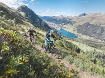 Zwei Biker fahren auf einem Singletrail in der Umgebung von Davos Klosters während man im Hintergrund den See sieht.