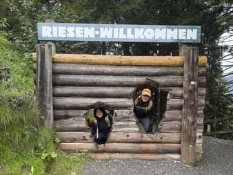 Zwei Wanderer mit Rucksäcken schauen durch Öffnungen in einer Holzstammstruktur mit 'Riesen-Willkommen'-Schild, umgeben von grünem Wald.