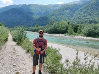 Ein Mann im Wanderoutfit inklusive Cap, Sonnenbrille und Wanderstöcken steht auf einem Wanderweg neben dem Fluss Soca, Slowenien. Im Hintergrund befinden sich bewaldete Berge.