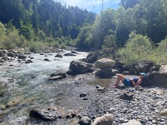Wanderer entspannt am steinigen Ufer neben einem rauschenden Bergbach. Bewaldete Hügel und blauer Himmel im Hintergrund bei Oey.