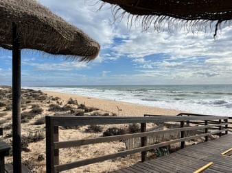 Holzsteg mit Strohdächern mit Blick auf Sandstrand und Meer im Naturpark Ria Formosa. Dünen mit Vegetation und Wellen unter bewölktem Himmel.