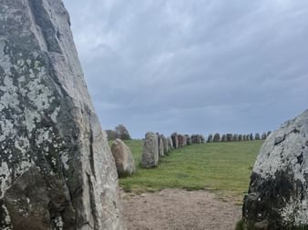 Blick durch flechtenbewachsene Steine bei Ales Stenar nahe Kåseberga. Die Steine bilden ein schiffsförmiges Muster auf grünem Gras unter bewölktem Himmel.