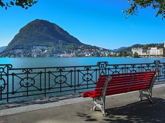 Banc de parc rouge sur la promenade du lac à Lugano avec balustrade en fer ornée, vue sur les eaux bleues du lac et toile de fond montagnarde.