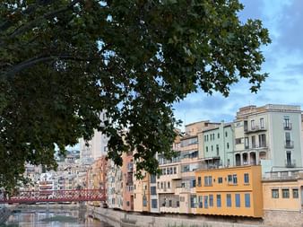 Bunte Häuser am Flussufer in Girona spiegeln sich im ruhigen Wasser, eingerahmt von grünen Baumzweigen. Eine rote Brücke ist im Hintergrund sichtbar.