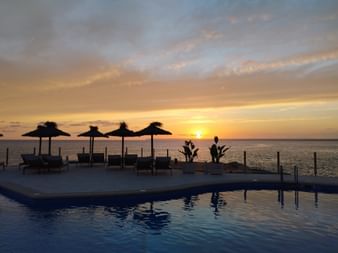 Pool mit Liegestühlen und Strohsonnenschirmen direkt am Meer bei Sonnenuntergang im Hotel Universal Cabo Blanco, Mallorca.
