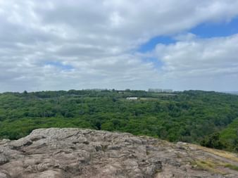 Felsformation im Vordergrund mit Blick auf grüne bewaldete Hügel und ein weißes Gebäude unter bewölktem Himmel bei Kivik am Österlenleden.
