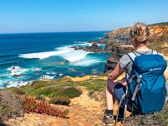 Wanderer mit blauem Rucksack sitzt auf Klippe mit Blick auf Atlantik am Rota Vicentina Weg. Dramatische Küste mit Wellen und Vegetation sichtbar.