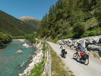 Zwei Velofahrer fahren der Inn im Graubünden entlang auf einem Kiesweg.