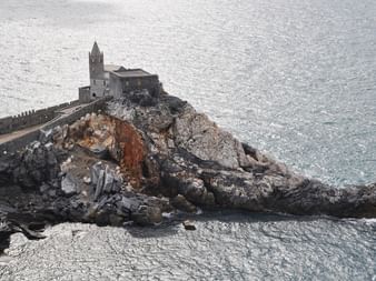 Steinkirche mit Glockenturm auf einem felsigen Küstenvorsprung, durch einen Gehweg mit dem Ufer verbunden, umgeben von ruhigem blauen Meer.