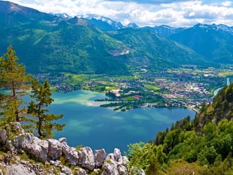 Vue panoramique du lac Traunsee entouré de montagnes vertes et de sommets alpins. Une ville se trouve le long du rivage avec des falaises rocheuses au premier plan.