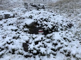 Rot-weißes Wegzeichen teilweise sichtbar zwischen schneebedeckten Büschen am Alpenpässeweg. Die alpine Landschaft ist mit frischem Schnee bedeckt.