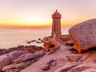 Phare de Ploumanac'h sur rochers de granit rose au coucher du soleil. La tour en pierre surplombe l'océan Atlantique avec un ciel orangé.