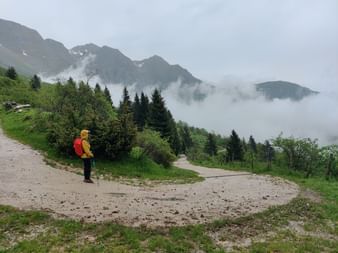 Eine Person wandert bei Regen und Nebel auf einem Wanderweg umgeben von Bäumen und Bergen.