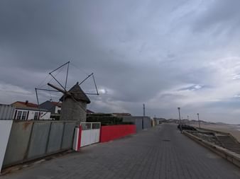 Traditionelle Steinwindmühle mit dreieckigen Segeln an der Küstenpromenade in Aguçadoura unter dramatischem Wolkenhimmel, Strand und Meer sichtbar.
