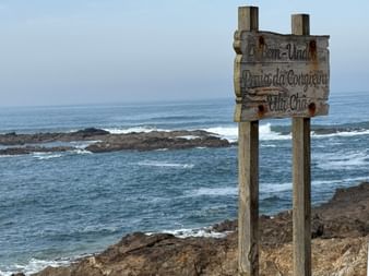 Verwittertes Holzschild mit Aufschrift 'Bem-Vindo Praia da Congreira Vila Chã' an felsiger Küste mit Atlantikwellen und Felsen im Hintergrund.