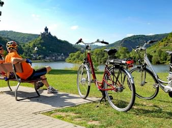 Deux cyclistes casqués assis sur un banc près de la Moselle avec leurs vélos garés à proximité. Un château médiéval se dresse sur une colline.