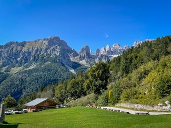 Brenta-Dolomiten mit gezackten Gipfeln unter blauem Himmel. Grüne Wiese mit Holzhütte und dichtem Wald im Vordergrund.
