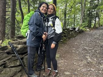 Zwei lächelnde Wanderinnen mit Rucksäcken und Wanderstöcken auf einem Waldweg. Steinmauer und grüne Bäume im Hintergrund sichtbar.
