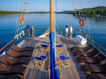 Sonnendeck der MS Macek mit braunen Liegen und blauen Stühlen. Passagiere stehen an der Reling und blicken auf das ruhige blaue Wasser und grüne Hügel.