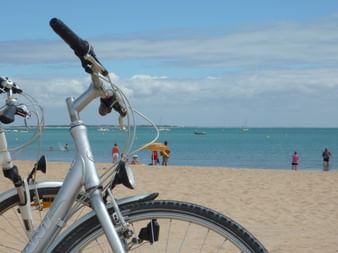 Deux vélos argentés garés sur une plage de sable avec océan turquoise et personnes en arrière-plan. Ciel bleu clair avec nuages blancs.