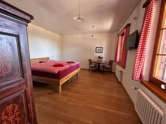 Helles Zimmer in der Gantrischhütte mit Holzbett, rot-weiß karierter Bettwäsche und Vorhängen, Holzschrank, Tisch mit Stühlen und Wandfernseher.
